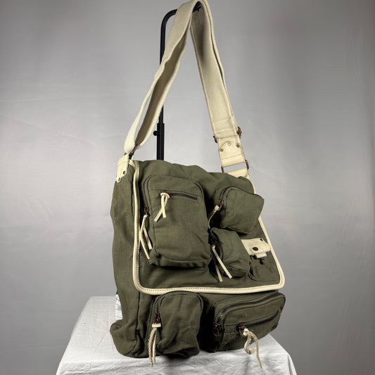 An olive green canvas crossbody bag with cream-colored trim and straps, displayed on a white draped surface against a gray studio background.