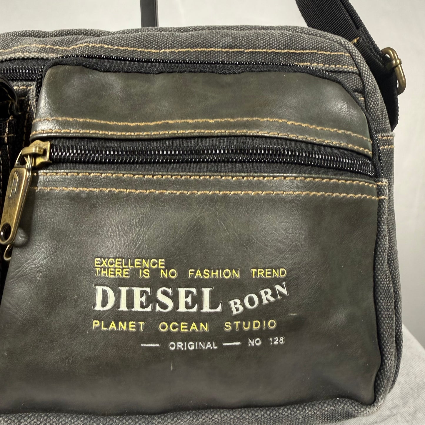 A close-up of a dark gray leather and canvas crossbody bag with gold stitching, a gold zipper pull, and a gold printed logo that reads 'DIESEL BORN PLANET OCEAN STUDIO'.
