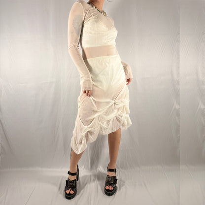 A model wearing a cream-colored two-piece outfit consisting of a long-sleeve crop top and a ruched midi skirt, paired with black platform sandals, set against a wrinkled white studio backdrop.