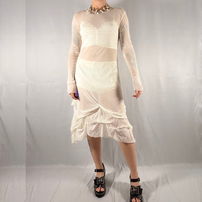 A model wearing a cream-colored sheer mesh long-sleeve dress with a ruffled hem, paired with black platform sandals and a chunky silver necklace, standing against a wrinkled light gray backdrop.