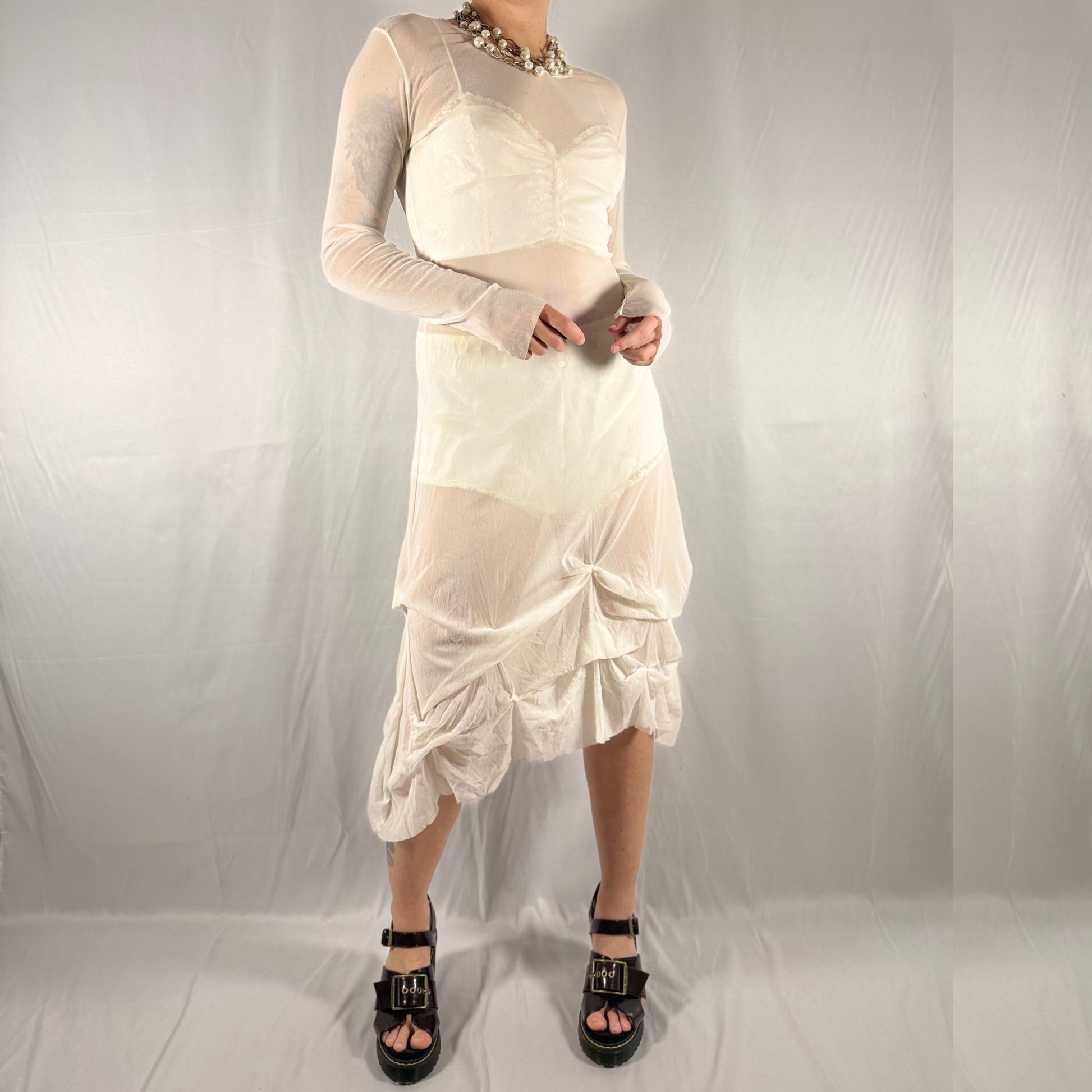 A model wearing a vintage-style off-white/cream dress with sheer mesh sleeves and a gathered, ruffled asymmetrical hem, paired with black platform sandals and a chunky beaded necklace, set against a wrinkled light gray studio backdrop.