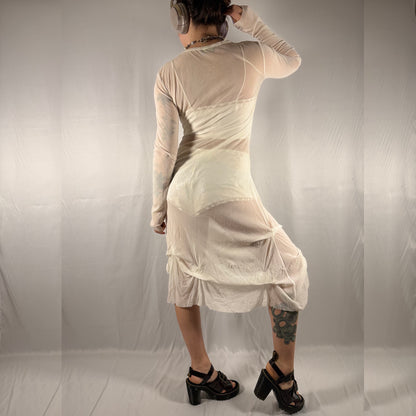 A person shown from the back wearing a sheer, long-sleeve, knee-length dress in a light cream or off-white color, paired with black high-heeled sandals, against a draped light gray fabric backdrop.