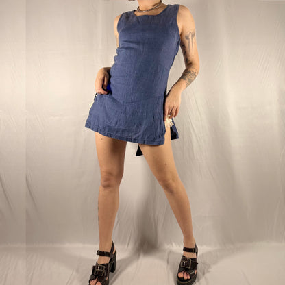 A person modeling a sleeveless blue denim mini dress against a wrinkled light gray backdrop, paired with black platform sandals.