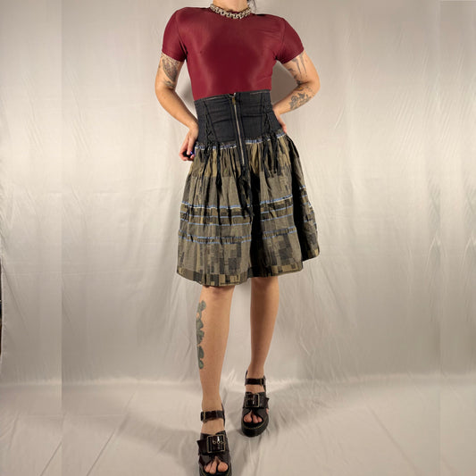 A model wearing a gothic punk style outfit consisting of a short-sleeve maroon top, a black corset-style waist, and a pleated plaid skirt in olive green, black, and gray. The model is standing against a wrinkled light gray fabric backdrop.