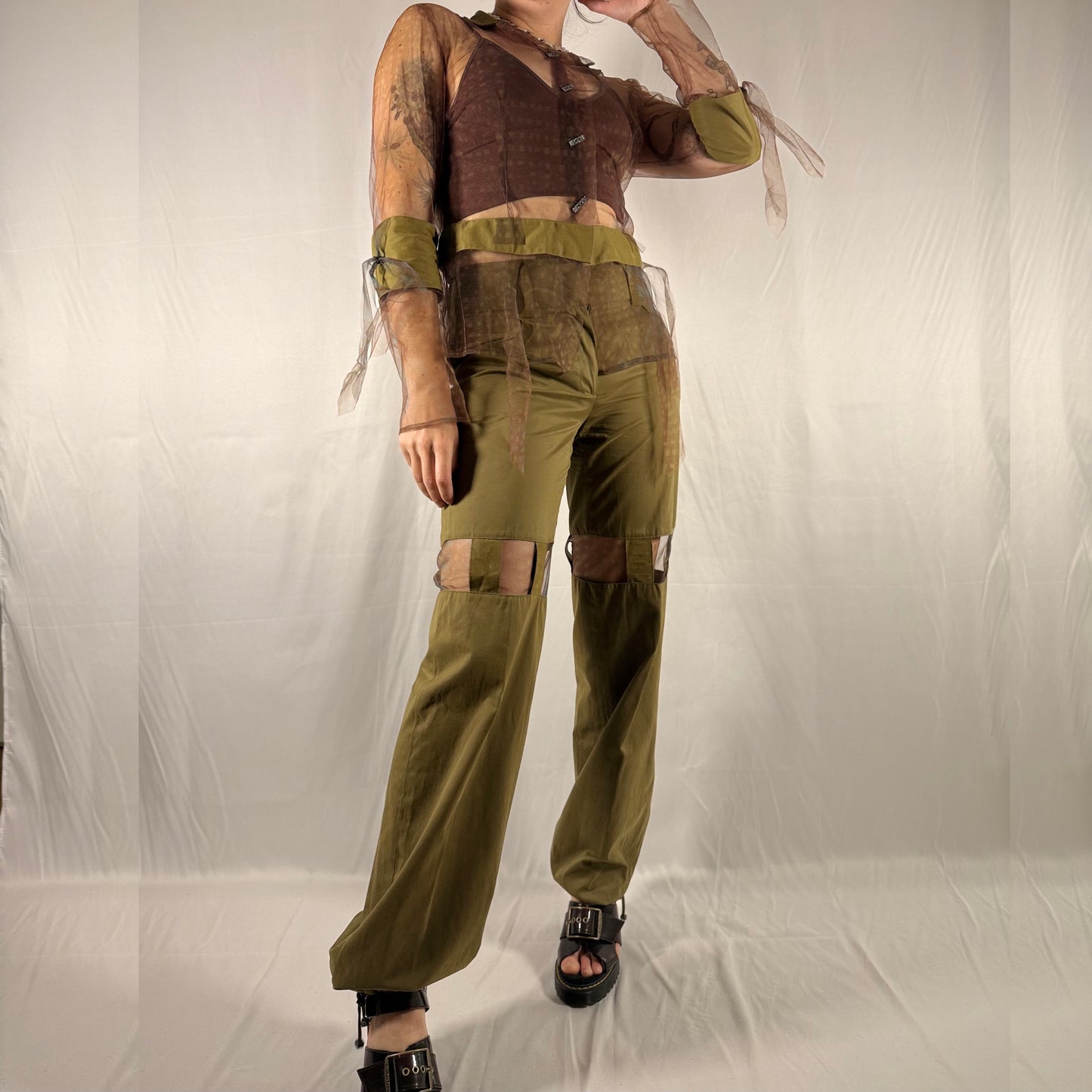 A person modeling a coordinated outfit consisting of olive green wide-leg pants with patchwork details, a matching olive green waistband and sleeve, and a brown cropped top with sheer sleeves, photographed against a wrinkled off-white studio backdrop.