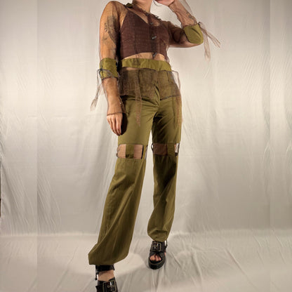 A person modeling a coordinated outfit consisting of olive green wide-leg pants with patchwork details, a matching olive green waistband and sleeve, and a brown cropped top with sheer sleeves, photographed against a wrinkled off-white studio backdrop.