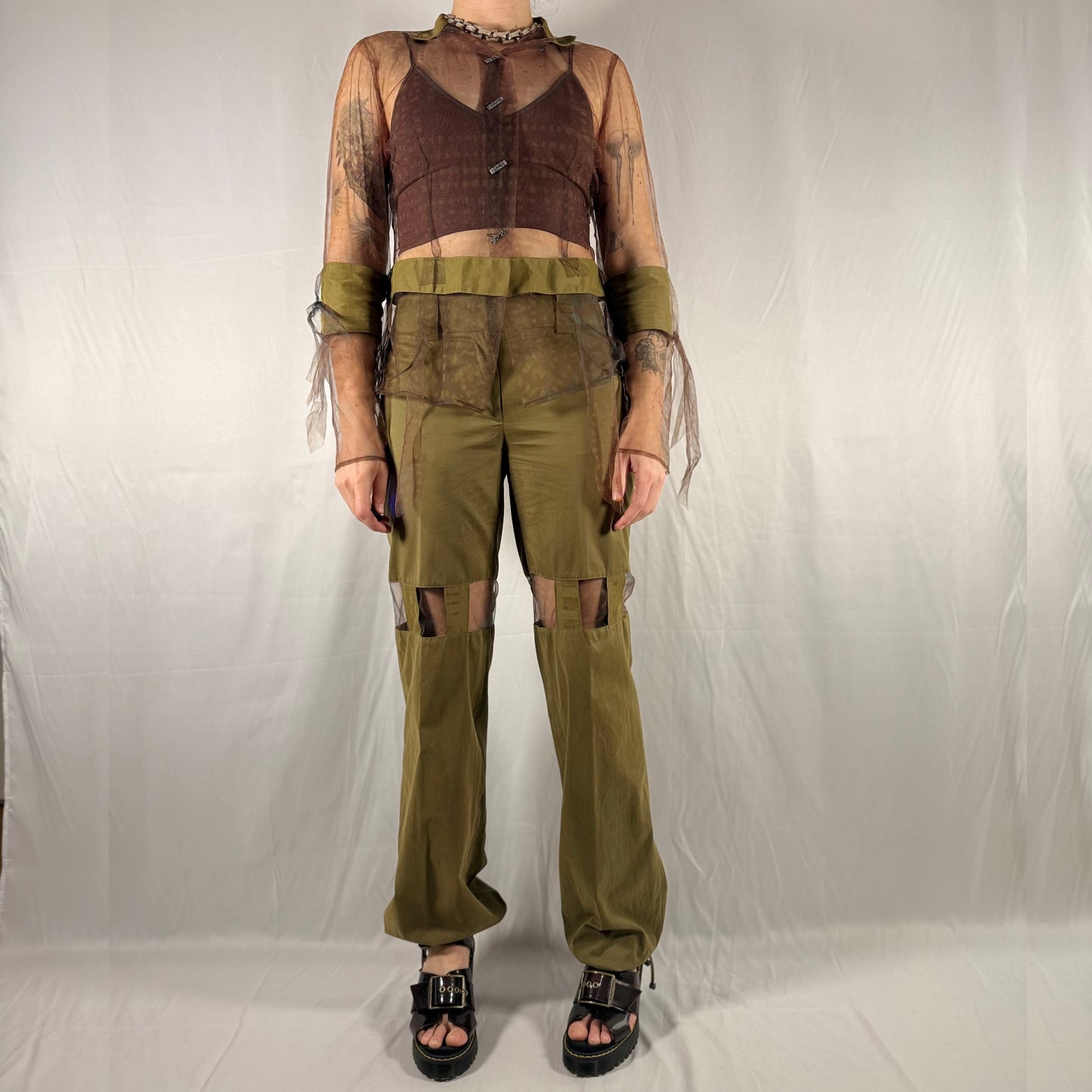 A model wearing a distressed, post-apocalyptic style outfit consisting of an olive green cargo pant leg with knee cutouts, a matching distressed green waist sash, a brown corset-style vest, and a sheer beige mesh long-sleeve crop top with fringe.