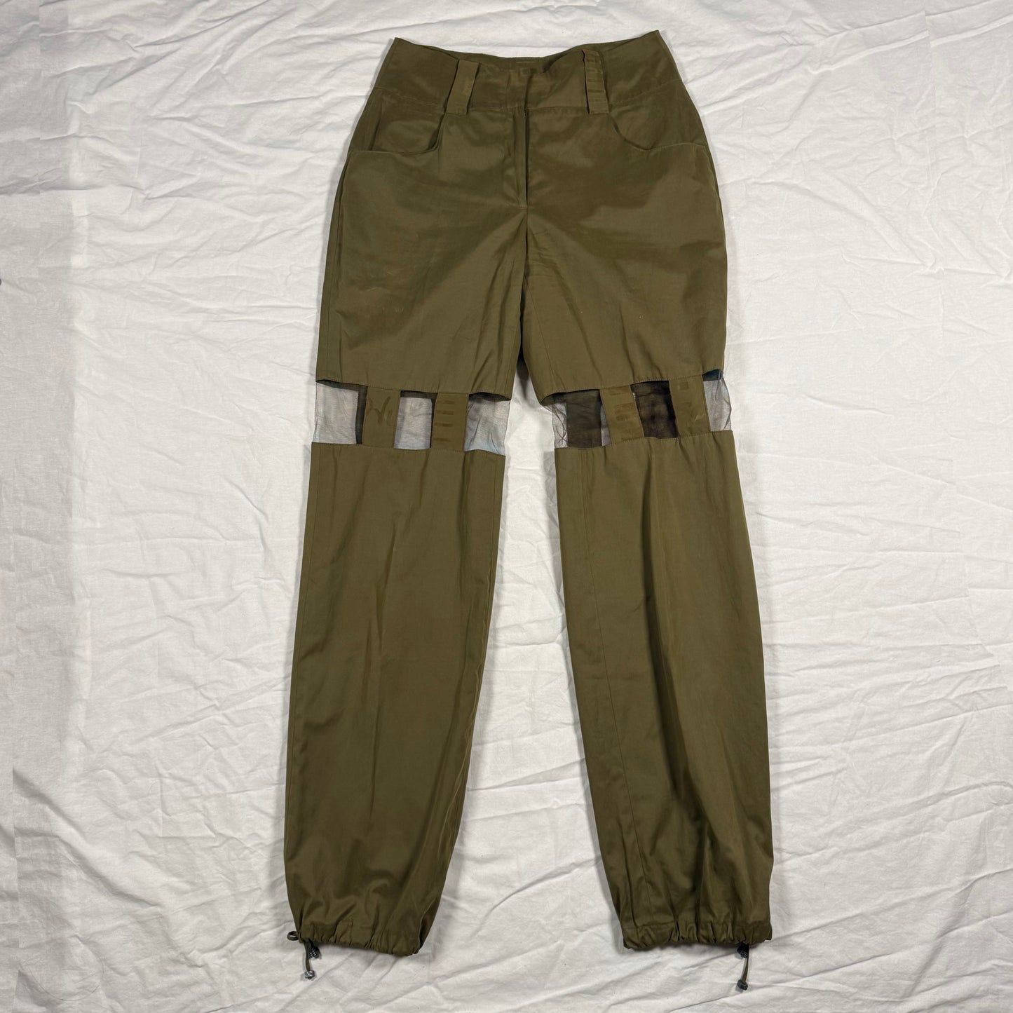 Olive green cargo pants with unique cutout details on the upper legs showing gray fabric lining, laid flat on a wrinkled white sheet.
