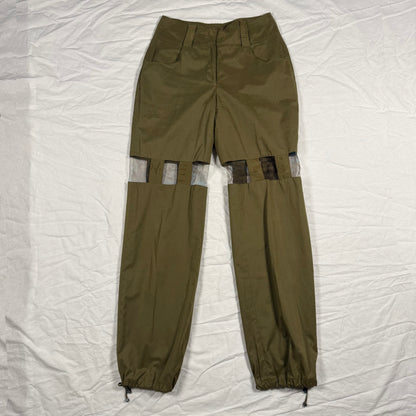 Olive green cargo pants with unique cutout details on the upper legs showing gray fabric lining, laid flat on a wrinkled white sheet.