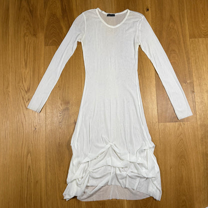 A white long-sleeve knit dress with a gathered, ruffled hem, laid flat on a light brown wooden floor.