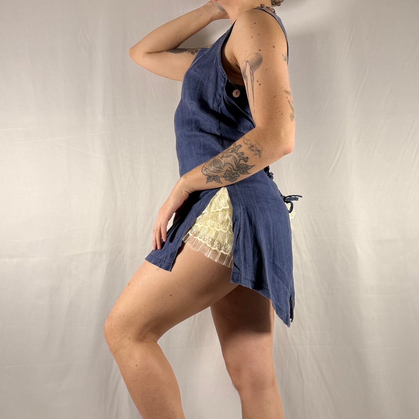 A woman modeling a sleeveless navy blue romper or short dress with a lace overlay, photographed against a light gray fabric backdrop.
