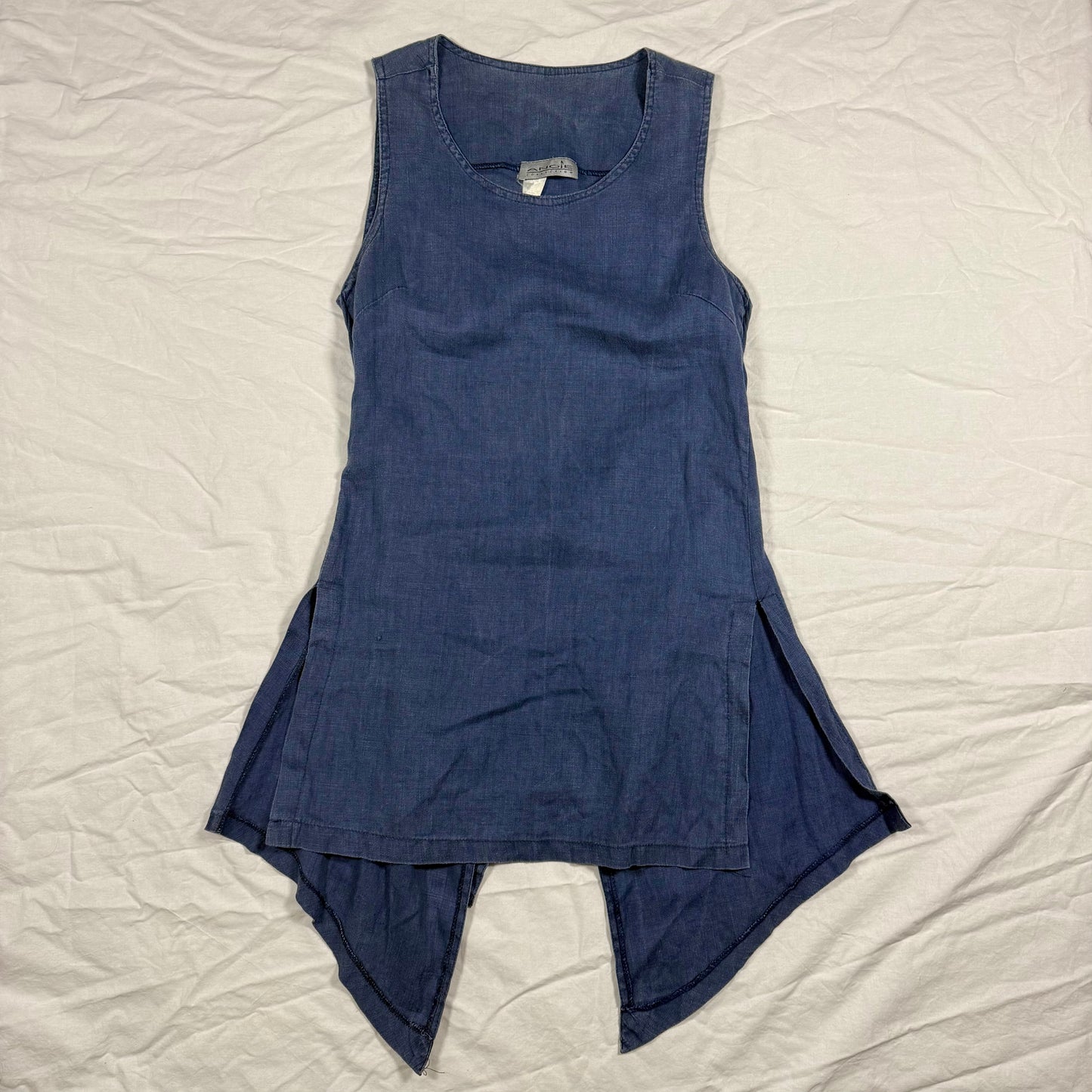 A sleeveless blue denim-like tunic dress with an asymmetrical hem, laid flat on a wrinkled white sheet.