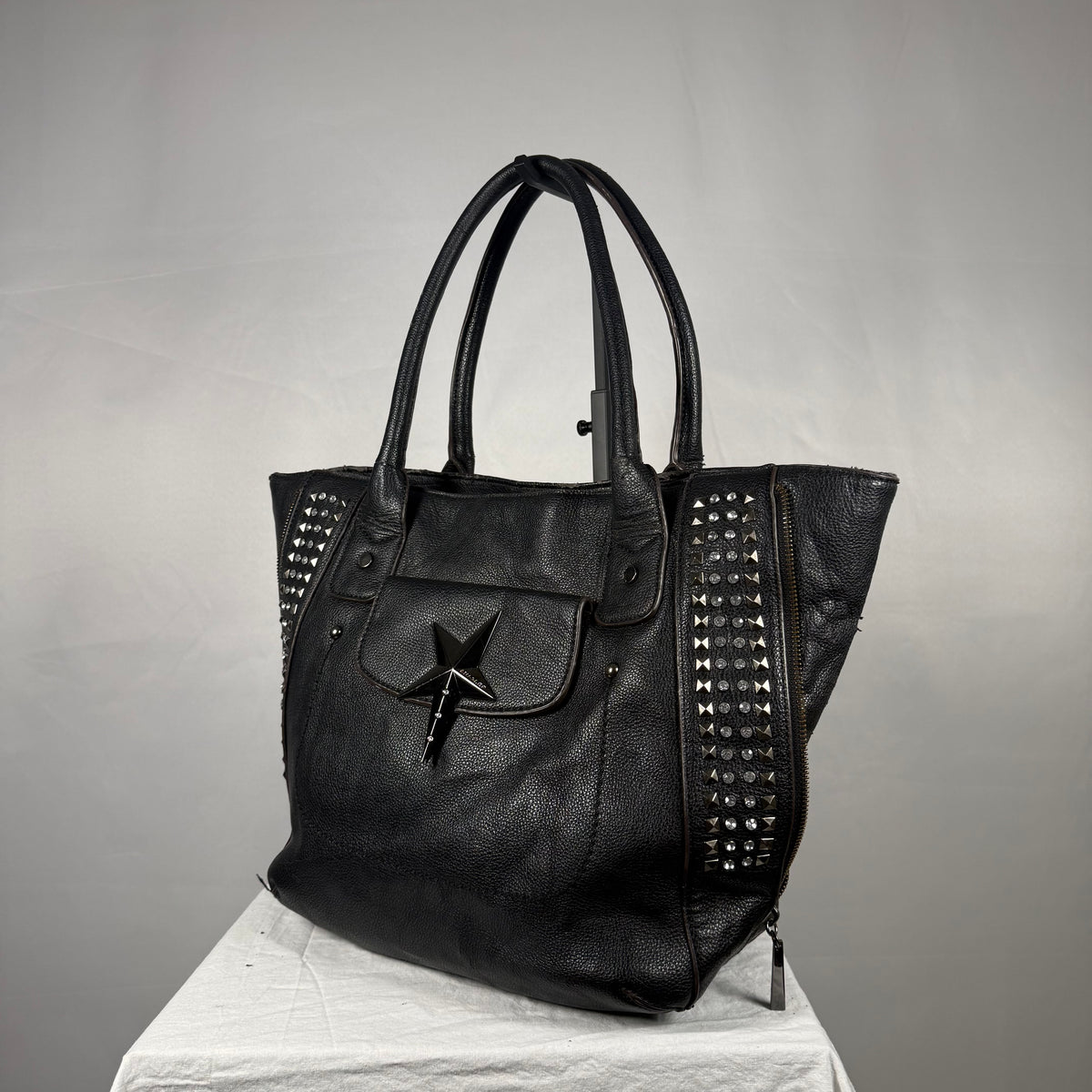 Black leather tote bag with silver studs on the sides, a front pocket with a star-shaped metal clasp, and dual top handles, displayed on a white draped surface against a gray background.