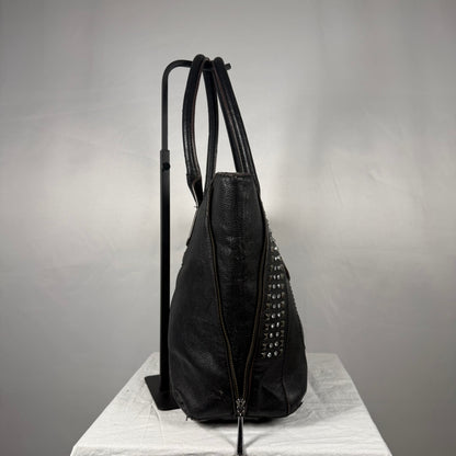 A black leather shoulder bag with silver studs, shown standing on a white cloth-covered surface next to a black display stand against a gray background.