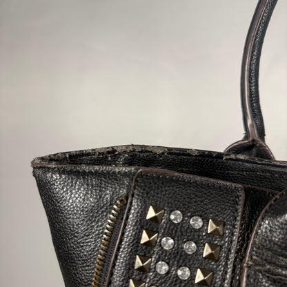 Close-up of a black pebbled leather handbag with gold-tone pyramid studs and clear rhinestones on a plain light gray background.