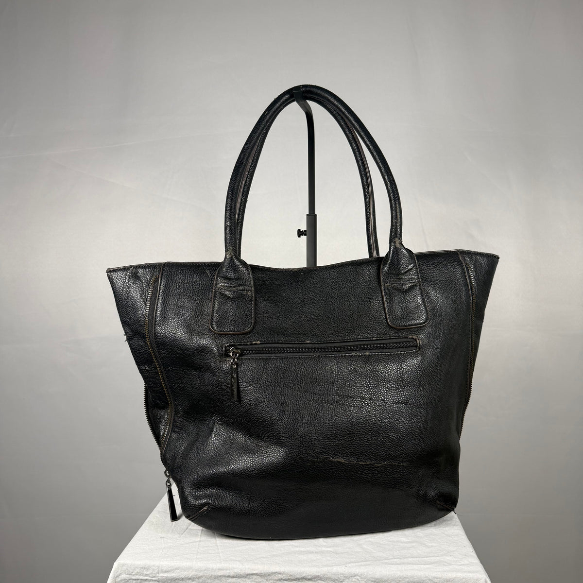 A black leather tote bag with dual top handles, a front zippered pocket, and side zipper details, displayed on a white fabric-covered stand against a gray studio background.