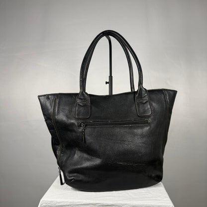 A black leather tote bag with dual top handles, a front zippered pocket, and side zipper details, displayed on a white fabric-covered stand against a gray studio background.