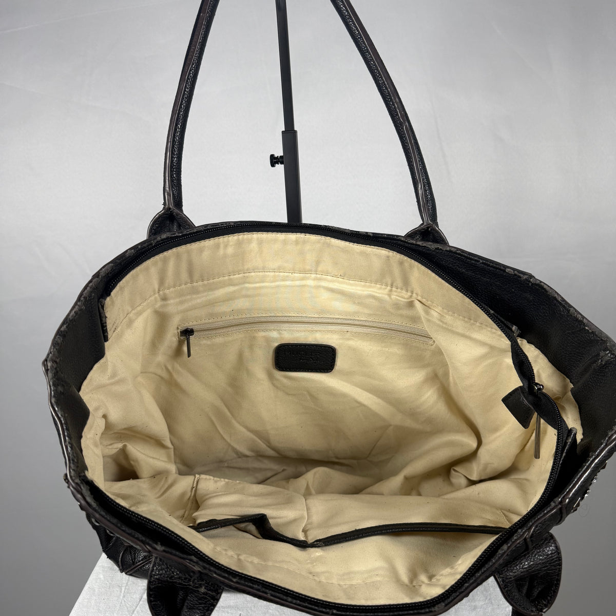 Black textured leather tote bag shown open from the front, revealing a cream-colored interior lining with a zippered pocket and black handles.