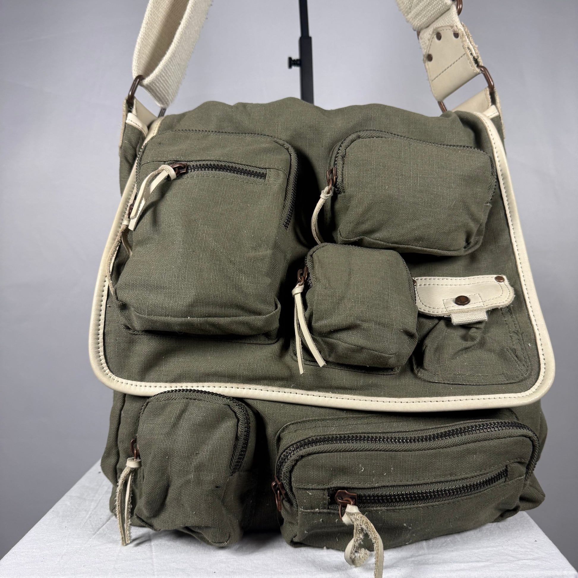 An olive green canvas shoulder bag with multiple utility pockets and cream-colored trim, displayed on a white surface against a gray background.