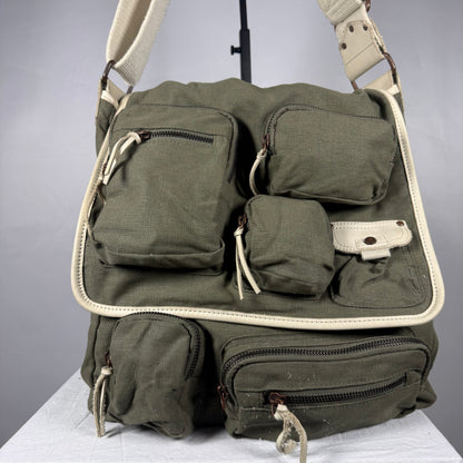 An olive green canvas shoulder bag with multiple utility pockets and cream-colored trim, displayed on a white surface against a gray background.