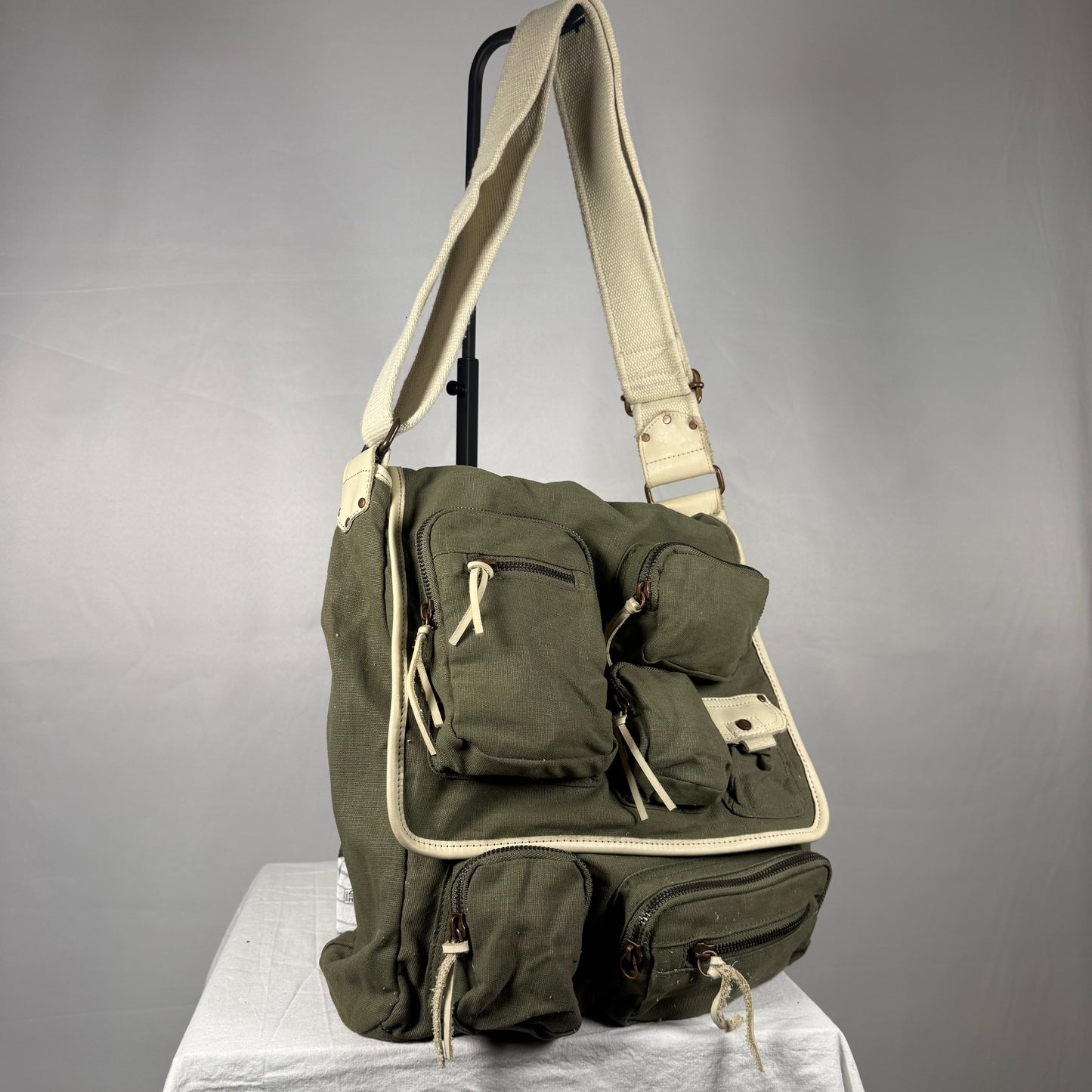 An olive green canvas crossbody bag with cream-colored trim and straps, displayed on a white draped surface against a gray studio background.
