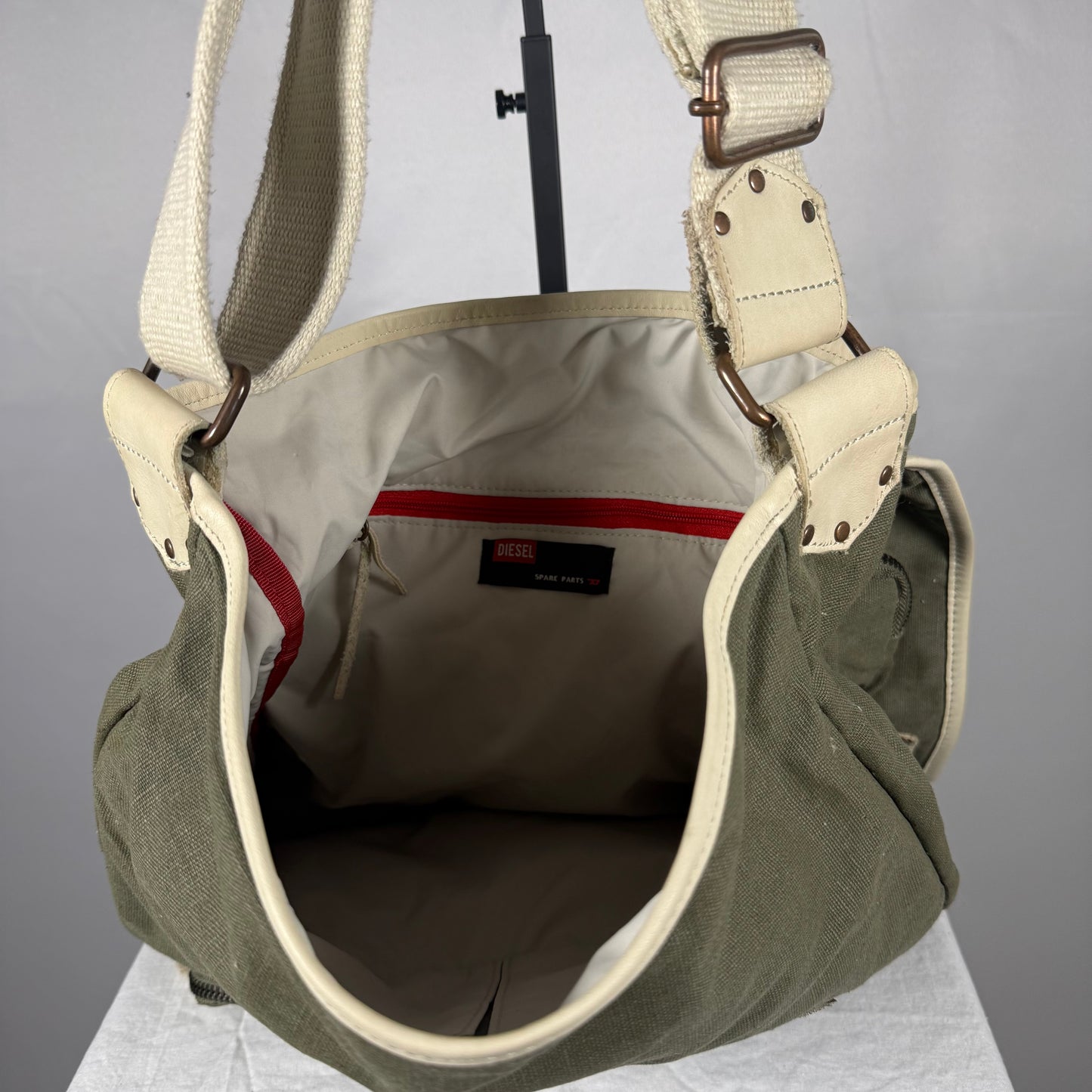 An olive green canvas shoulder bag with cream-colored webbing straps and metal hardware, shown open to display its interior lining and a red pocket detail.