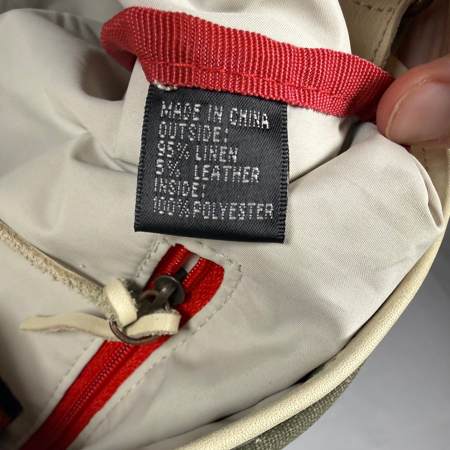 Close-up of a beige fabric bag with a red zipper and a black sewn-in label reading 'MADE IN CHINA 95% LINEN 5% LEATHER INSIDE 100% POLYESTER'. A finger is holding the bag open.