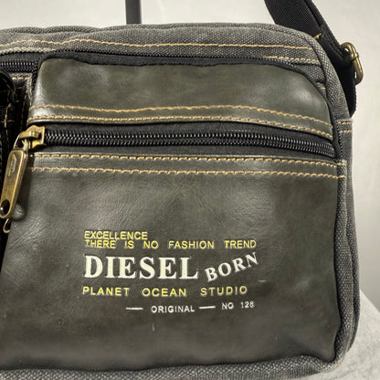 A close-up of a dark gray leather and canvas crossbody bag with gold stitching, a gold zipper pull, and a gold printed logo that reads 'DIESEL BORN PLANET OCEAN STUDIO'.