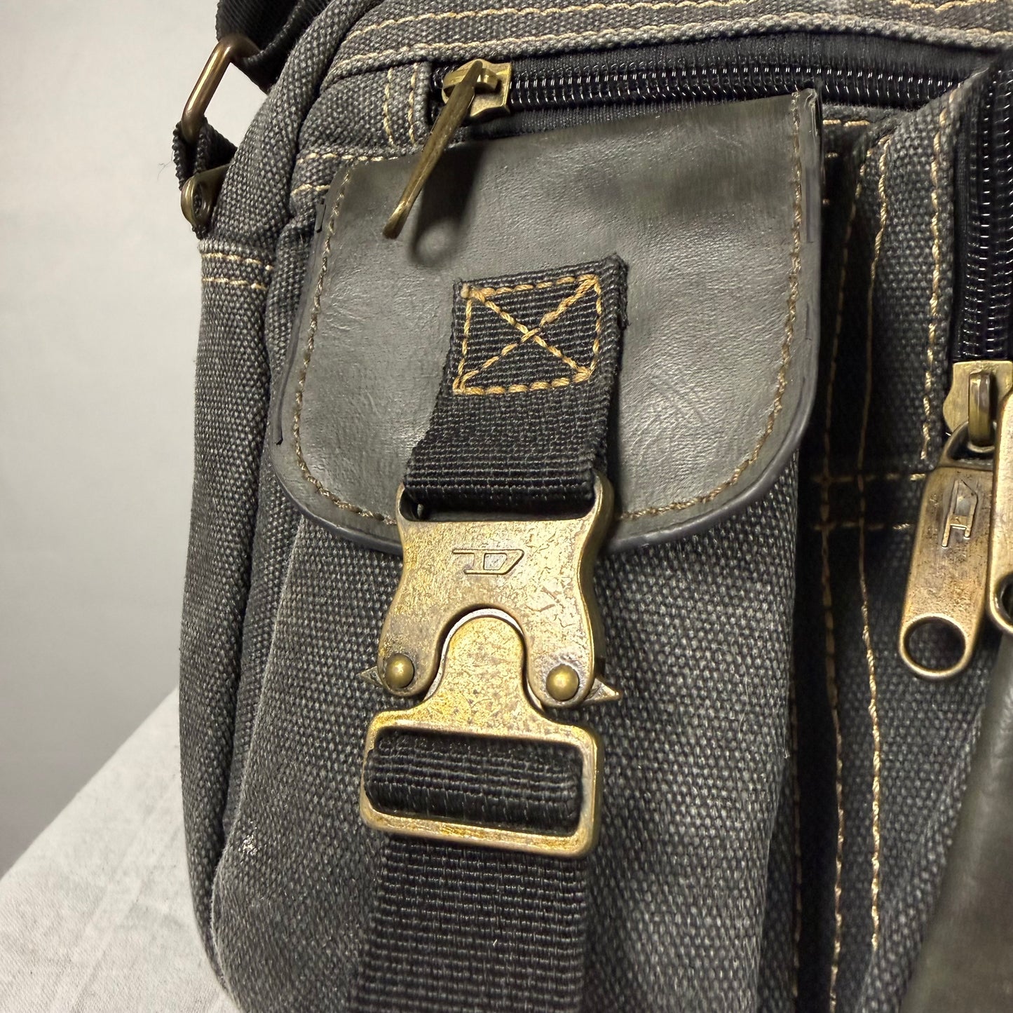 Close-up of a dark gray canvas and leather crossbody bag with an antique bronze buckle closure and zipper pulls.