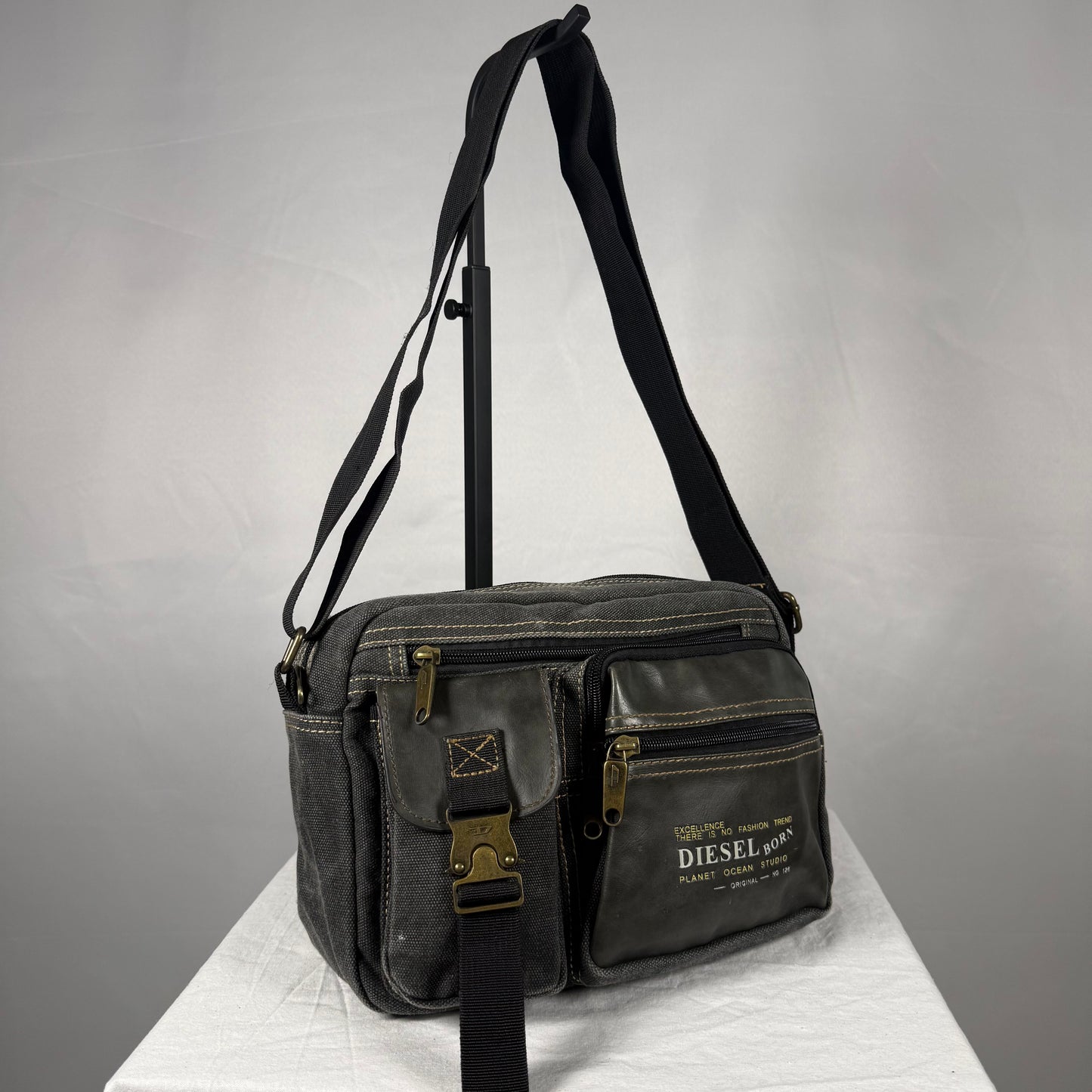 A dark gray canvas and leather crossbody bag with multiple zippered pockets and a black shoulder strap, displayed on a white pedestal against a gray background. The front pocket features brass hardware and a white printed logo that reads 'DIESEL WORKS'.