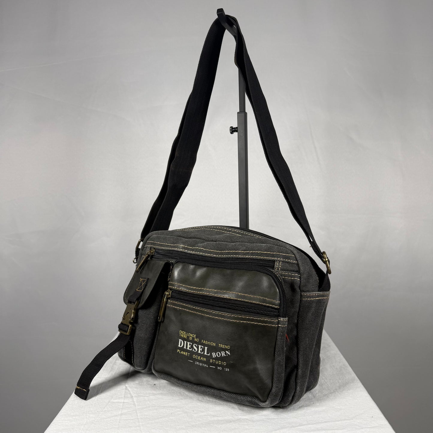 A black canvas crossbody bag with a front zippered pocket and a white printed 'DIESEL BORN' logo, displayed on a white surface against a gray background.