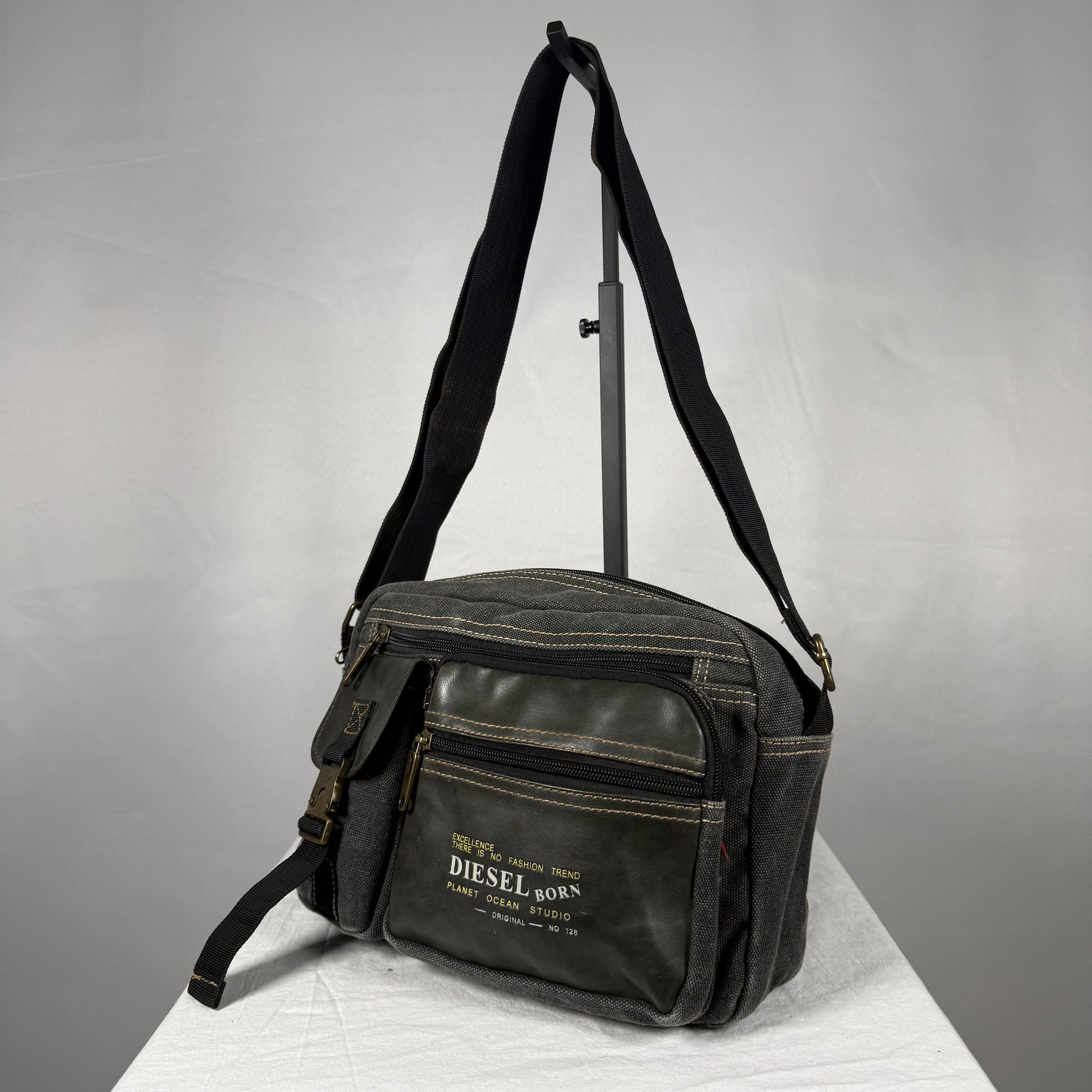 A black canvas crossbody bag with a front zippered pocket and a white printed 'DIESEL BORN' logo, displayed on a white surface against a gray background.