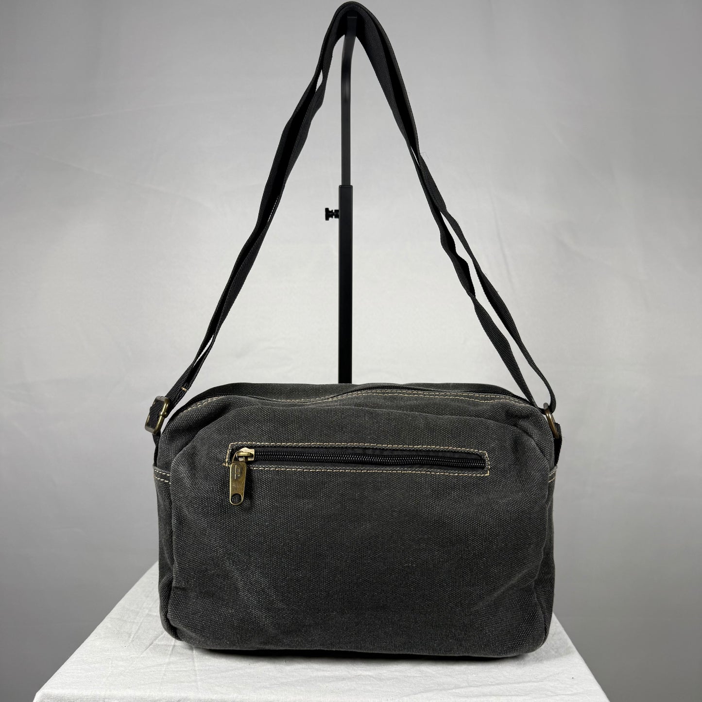 A dark gray canvas shoulder bag with a front zippered pocket and a gold-tone zipper pull, displayed on a white surface against a gray background.