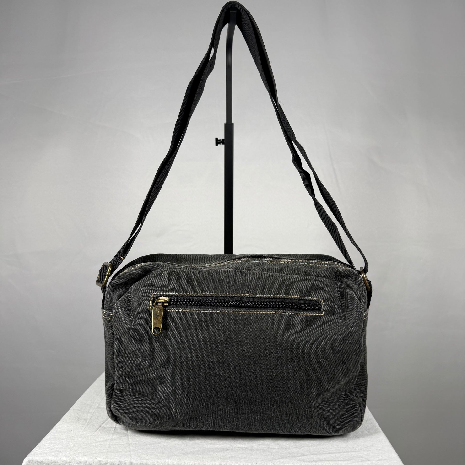 A dark gray canvas shoulder bag with a front zippered pocket and a gold-tone zipper pull, displayed on a white surface against a gray background.