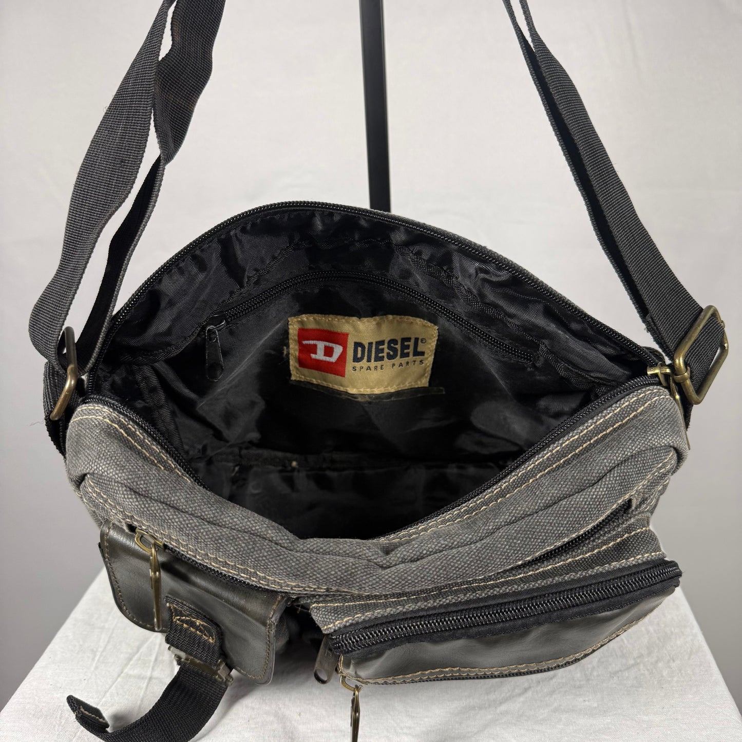 A Diesel crossbody bag shown open, featuring a gray canvas exterior, black interior lining, and brass hardware. The Diesel brand label is visible inside the bag.