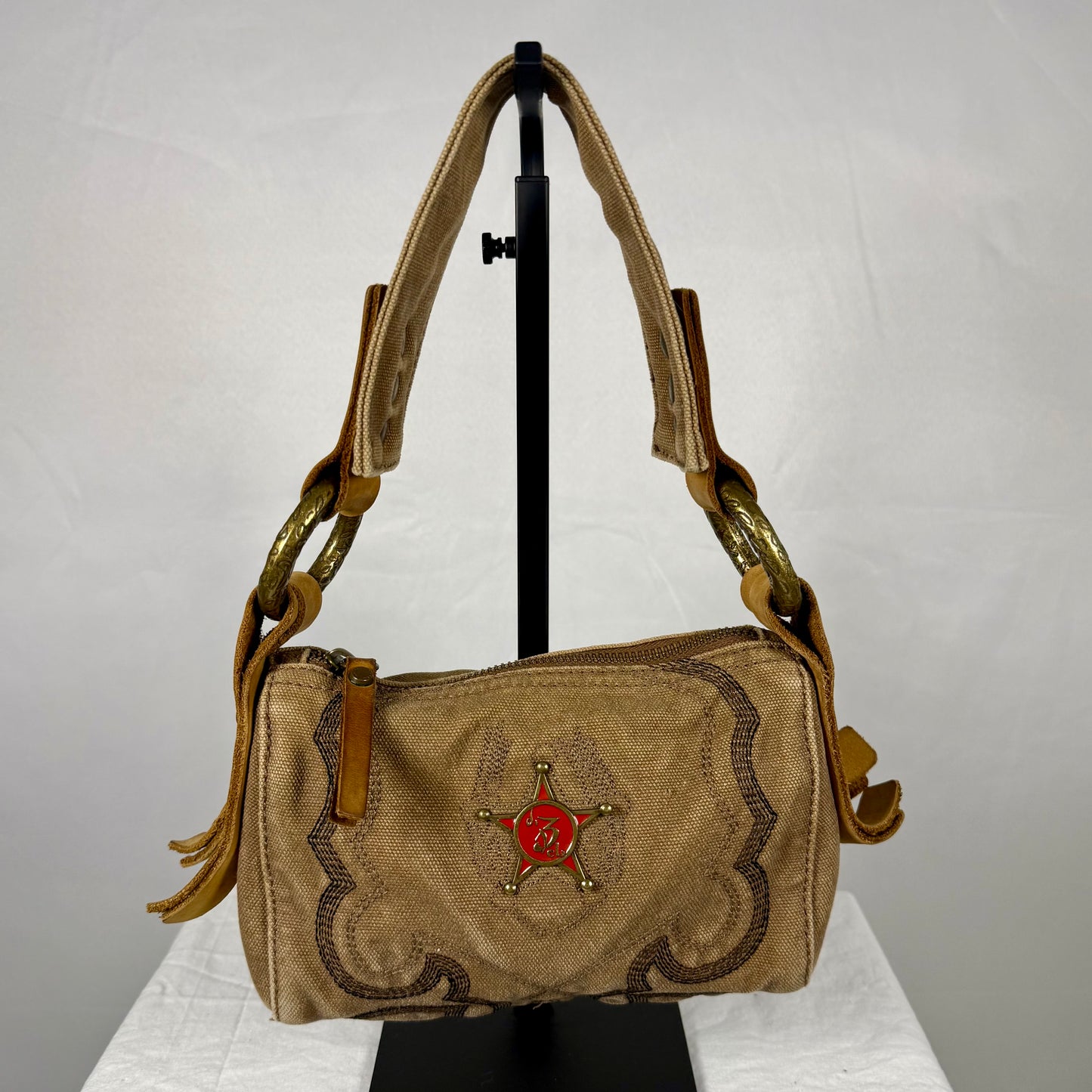 A vintage-style tan suede shoulder bag with brown embroidered scrollwork, gold-tone hardware, and a red sheriff's badge emblem on the front, displayed on a black stand against a white backdrop.