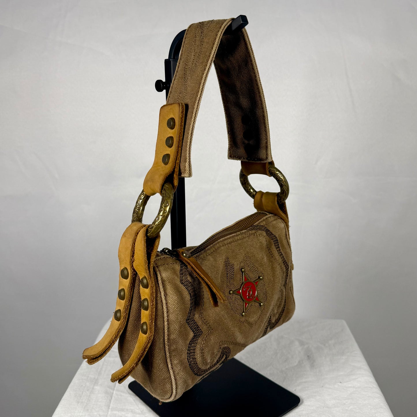 A vintage-style shoulder bag in tan suede and brown leather with a red star emblem, displayed on a black stand against a white backdrop.