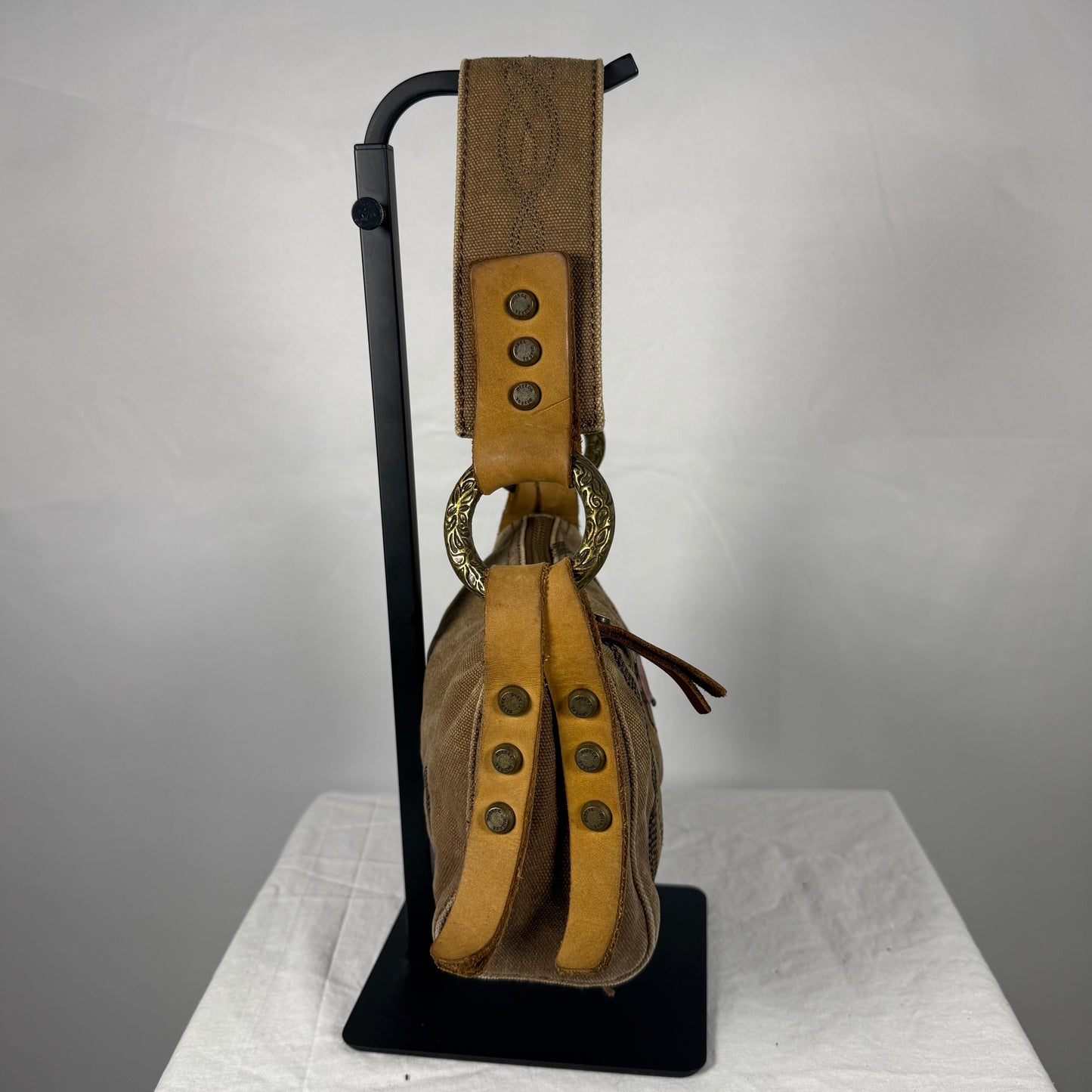 A brown leather shoulder bag with tan leather straps and brass-tone hardware, displayed on a black stand against a light gray background.
