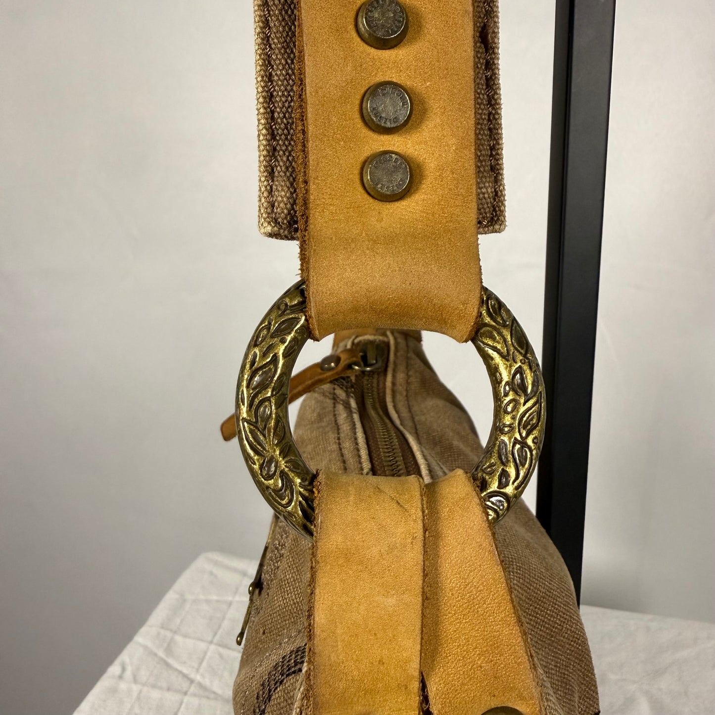 Close-up of a vintage-style shoulder bag featuring a tan suede strap with three decorative metal buttons, a large ornate gold-tone ring, and a beige woven fabric body.
