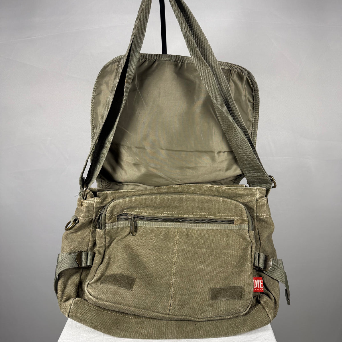 An olive green canvas crossbody bag with multiple front pockets and a top handle, shown open against a neutral gray background.