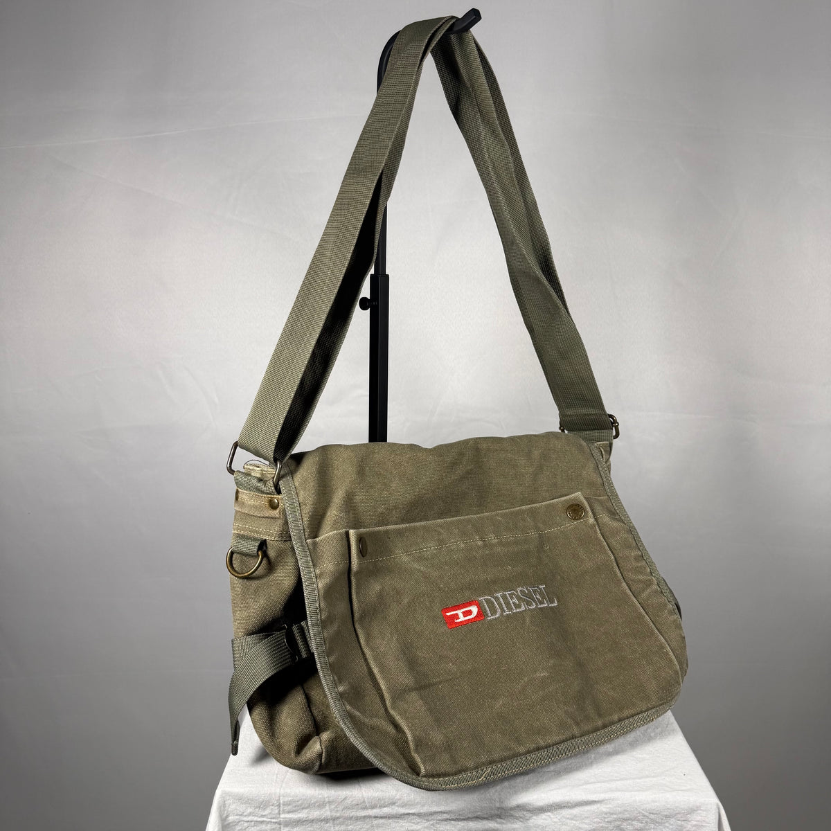 An olive green canvas shoulder bag with a front pocket featuring a red and white Diesel logo, displayed on a stand against a gray background.
