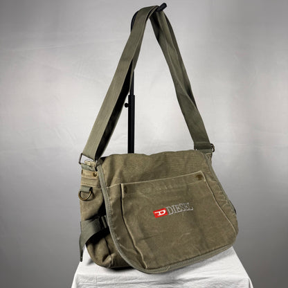 An olive green canvas shoulder bag with a front pocket featuring a red and white Diesel logo, displayed on a stand against a gray background.