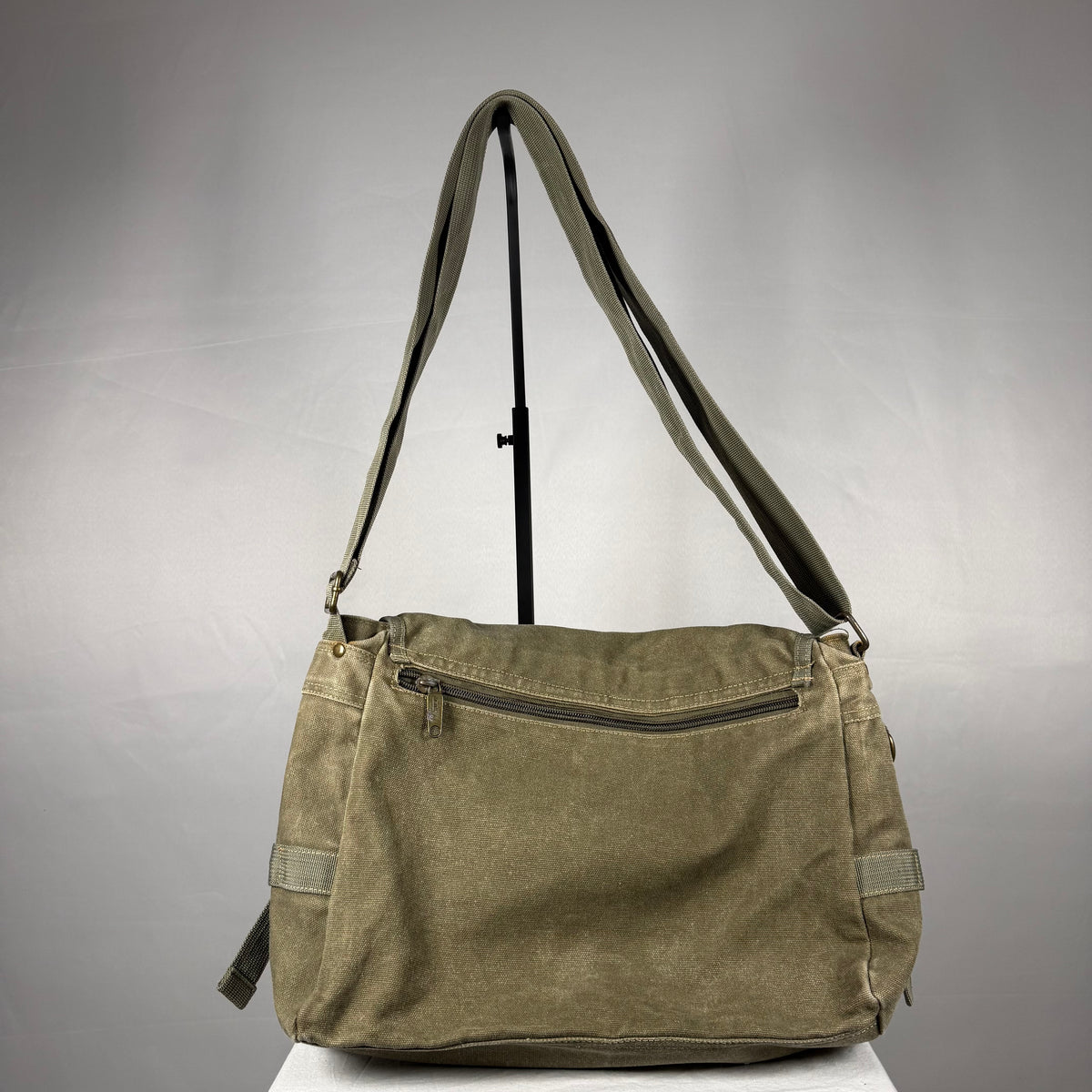An olive green canvas shoulder bag with a long adjustable strap, displayed on a stand against a neutral gray background.
