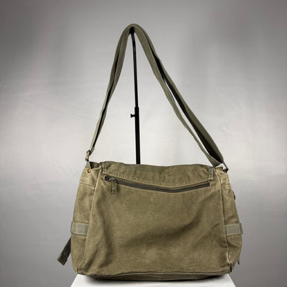 An olive green canvas shoulder bag with a long adjustable strap, displayed on a stand against a neutral gray background.