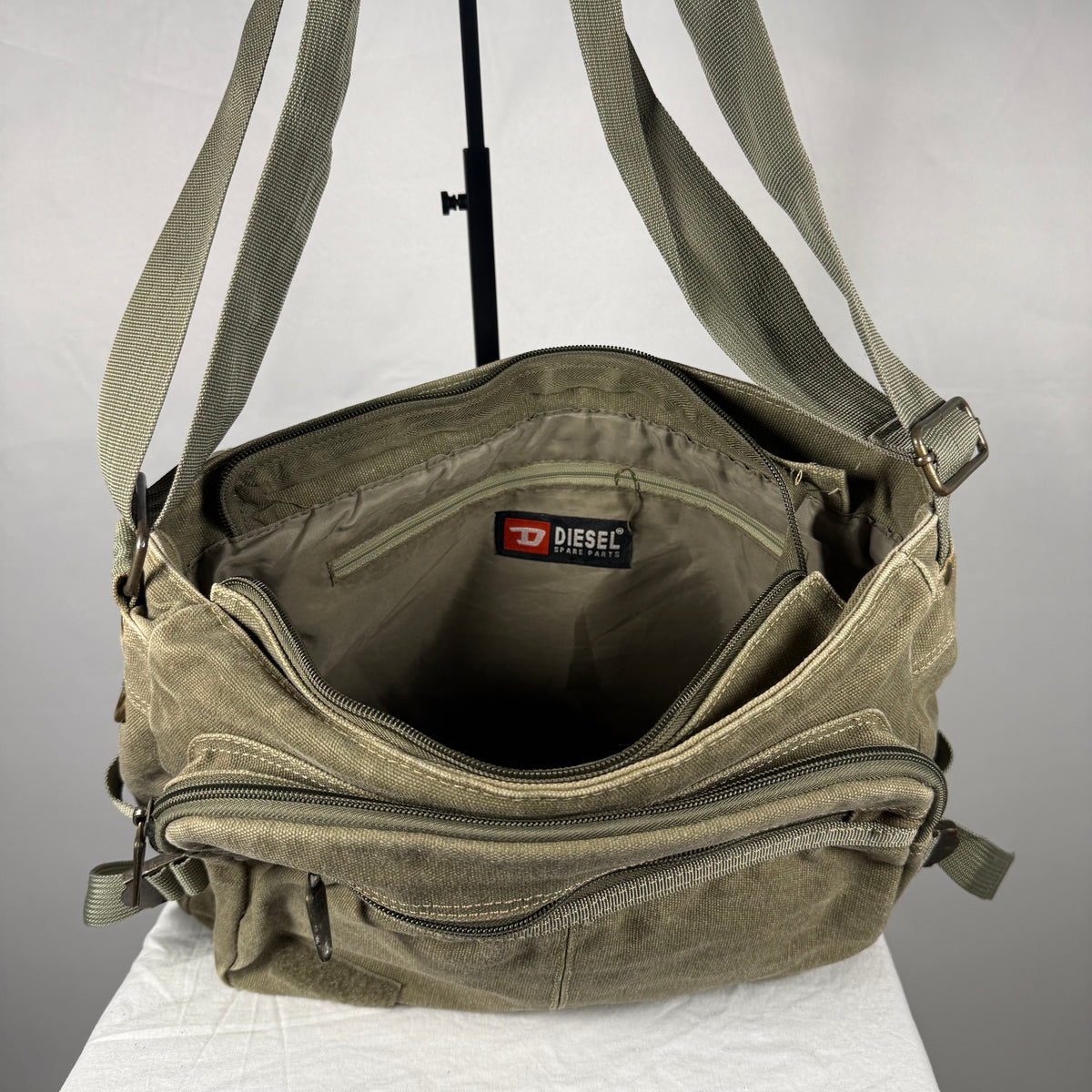 An olive green canvas crossbody bag by Diesel, shown open to display its interior compartments, including a zippered pocket and the Diesel brand label inside.