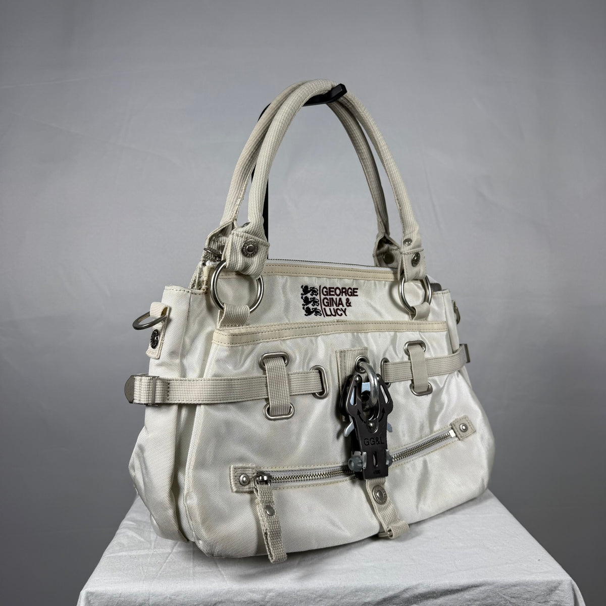 A light beige nylon handbag by George Gina & Lucy, featuring multiple straps, buckles, zippers, and a black horse-shaped charm, displayed on a white draped surface against a gray background.