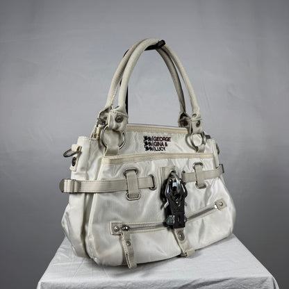 A light beige nylon handbag by George Gina & Lucy, featuring multiple straps, buckles, zippers, and a black horse-shaped charm, displayed on a white draped surface against a gray background.