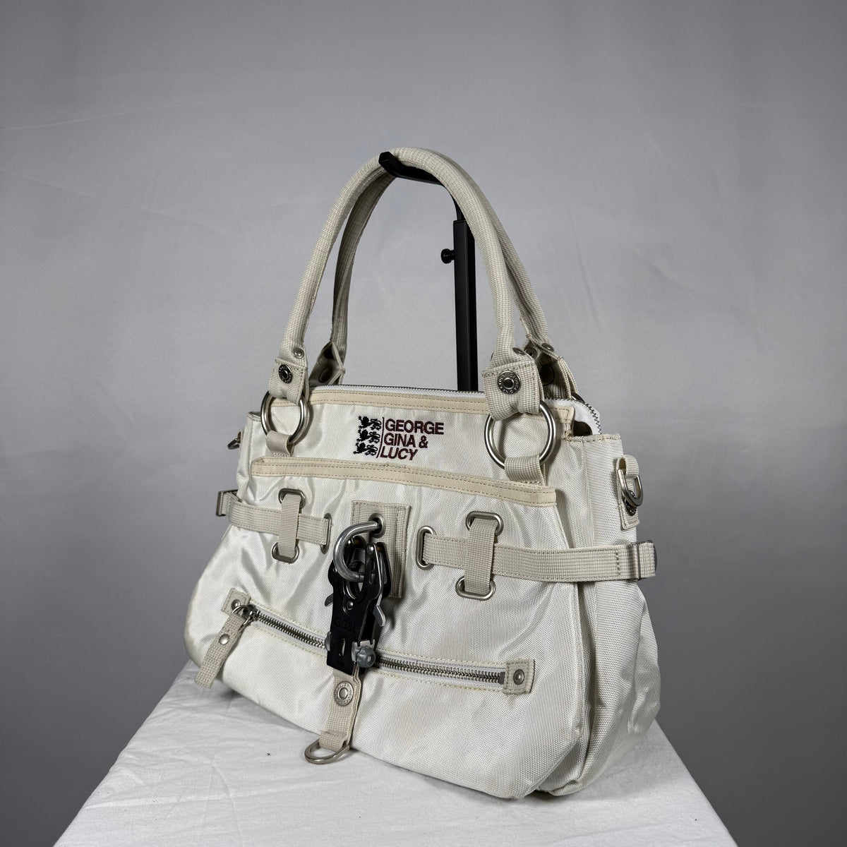 A vintage-style white nylon handbag with beige straps, silver-tone hardware, and a black embroidered patch reading 'GEORGE GINA & LUCY' on the front, displayed on a white surface against a gray background.