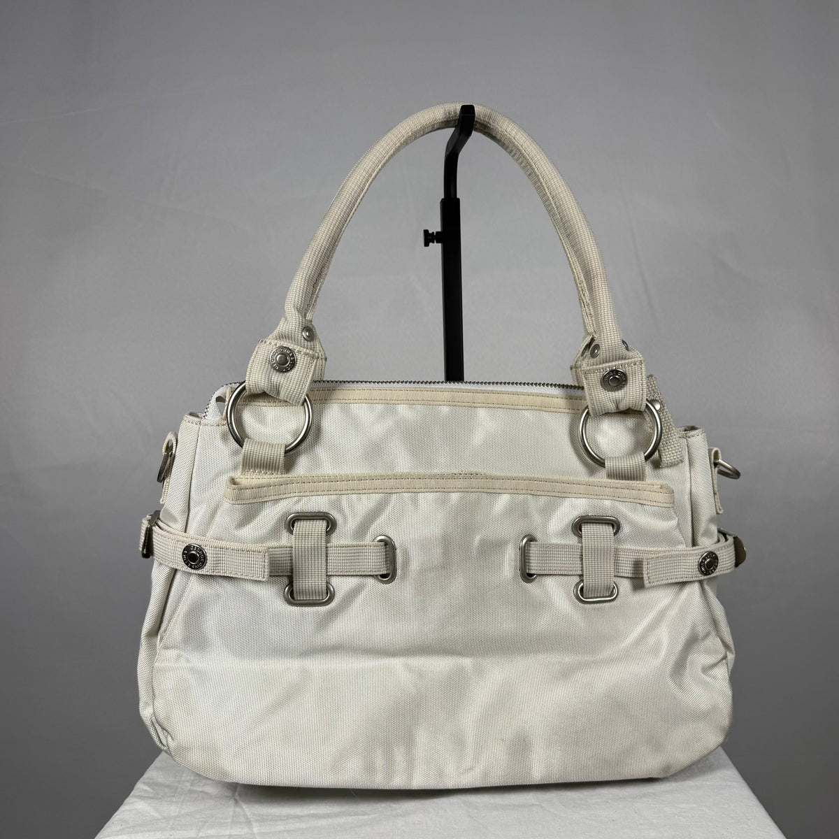 A light beige nylon handbag with silver-tone hardware, displayed on a black stand against a gray background.