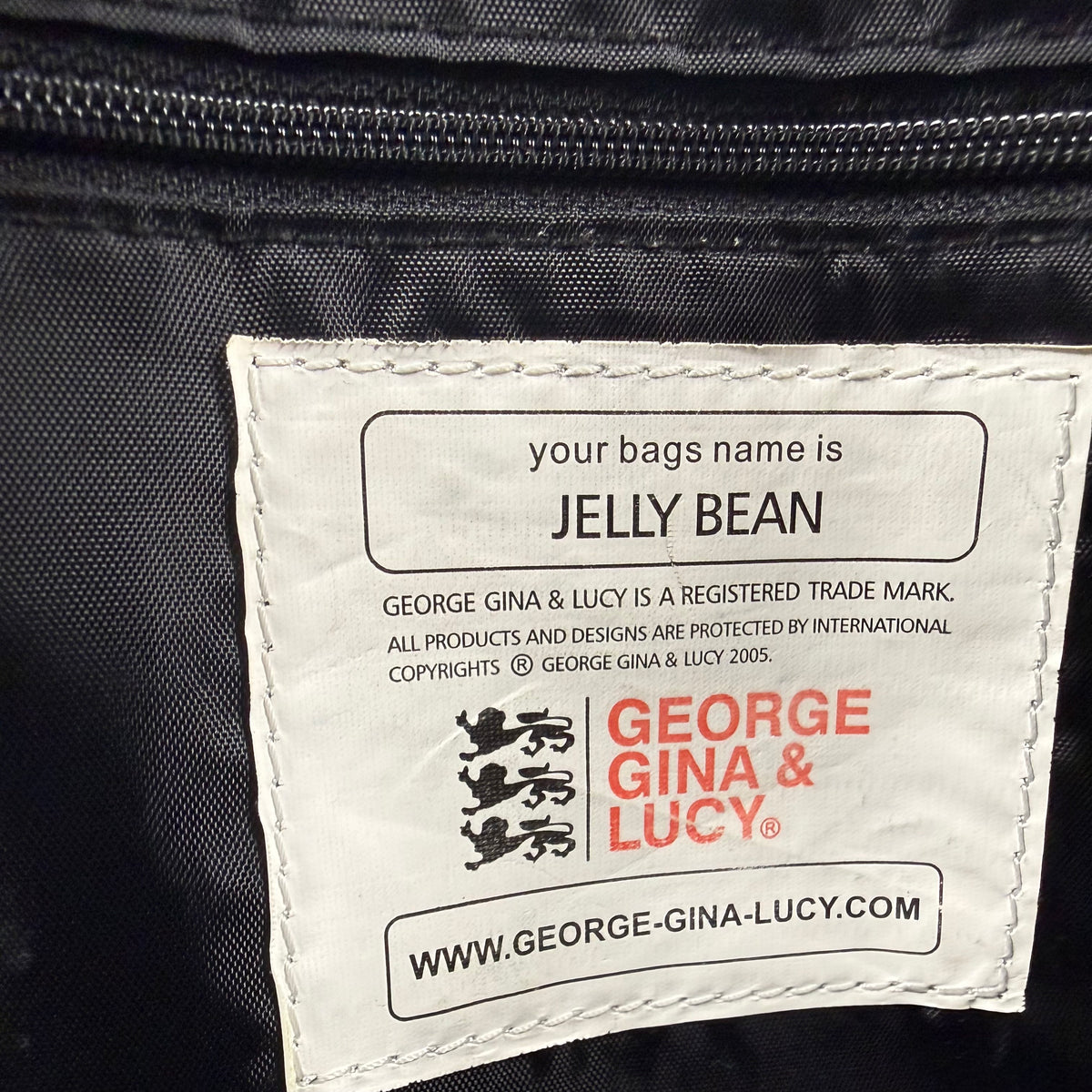 Close-up of a white sewn-in label on a black nylon bag, printed with the George Gina & Lucy brand name, website, and a customizable text field that reads 'your bags name is JELLY BEAN'.